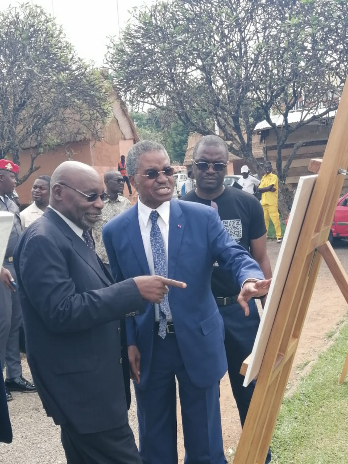 André Mama Fouda, en costume et lunettes sombres, devant un panneau d’exposition